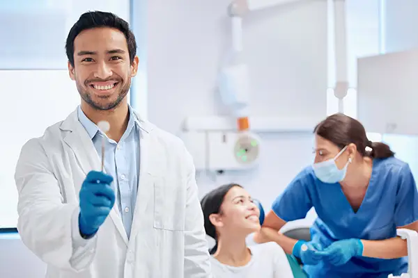 A smiling dentist holding an instrument while a patient and a dental assistant interact in the background during a dental check-up.